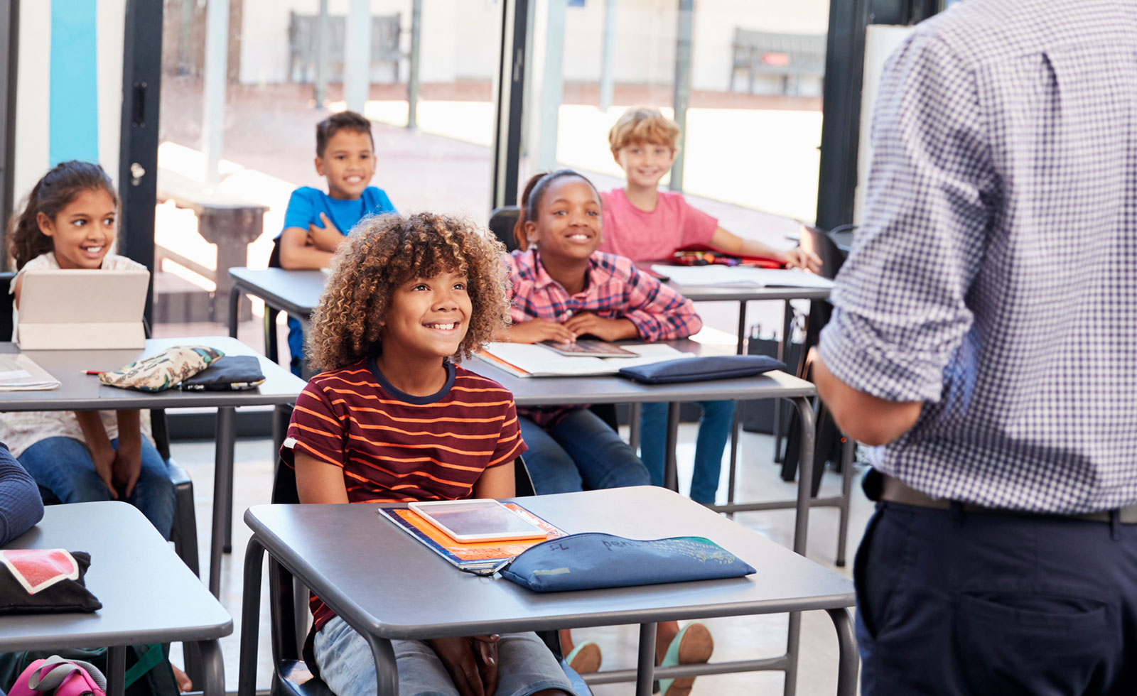 Elementary school students seated at desks in classroom, looking at teacher while smiling.