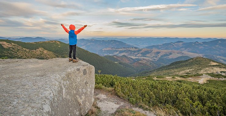 young person standing on a rock outcropping with their arms up looking out at mountains in the distance