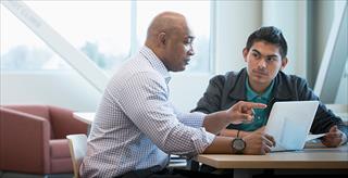 younger person being mentored by an older person sitting at a desk with a computer