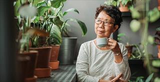 Older woman in glasses sitting by a window looking out surrounded by indoor plants and holding a teacup