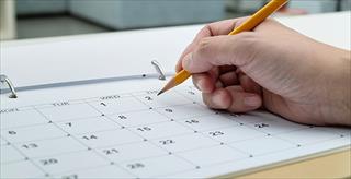 Hand holds pencil over calendar placed on desk.